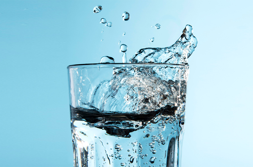 Glass of water with a dramatic splash rising above the rim, against a light blue background, capturing motion of pouring water.